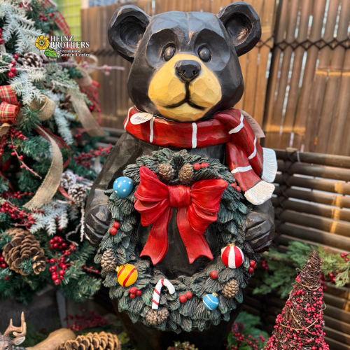 large, festive bear statue with holiday wreath in a decorative display with pinecones, glitter, tree surrounded by Christmas decorations.